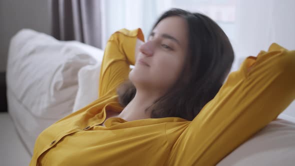 Young Girl Sits on the Sofa Raising Her Hands Behind Her Head in a Yellow Shirt Window Background