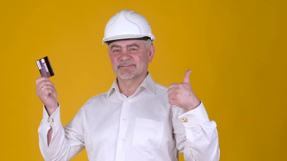 Cheerful Senior man builder in helmet hold in hand bank credit card showing thumbs up