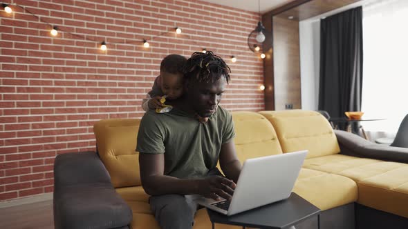 Smiling African Father Doing Freelance Work on Laptop While His Son Disturbing Him From the Back alt