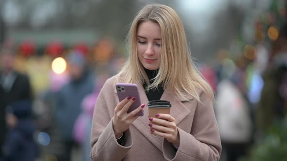 Lady with a cup of hot coffee uses Mobile Phone while standing on the street in the city. alt