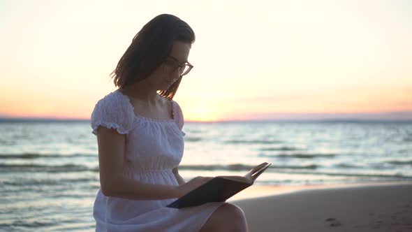 A Young Woman Sits on a Stone on the Beach By the Sea with a Book in Her Hands. A Girl in a White alt