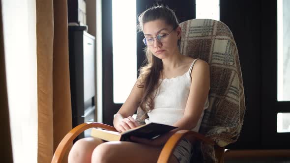 Young girl in glasses reads a book in natural light on a rocking chair alt