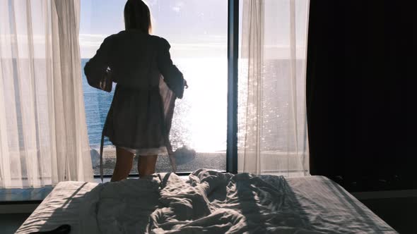 A Woman Puts on a Robe Looking Out the Panoramic Window Overlooking the Sea on a Sunny Day