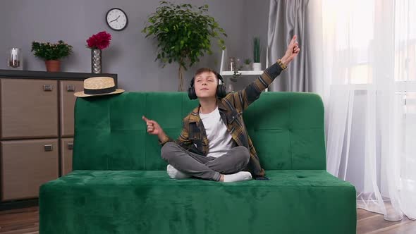 Boy Sitting on Sofa at Home and Enjoying Inflammatory Music in Headphones with Raised Hands alt