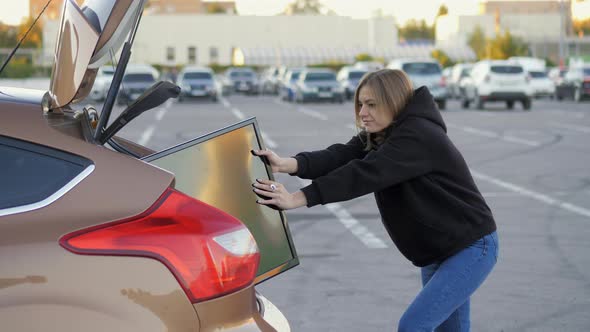 Woman Funny Trying to Put Purchased Modern Tv in Car Trunk at Supermarket Parking alt