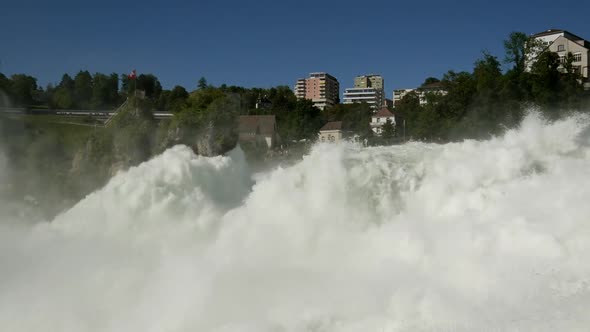 Zoom in shot of splashing rhine water fall with swiss flag on mountain and riding train in backgroun alt