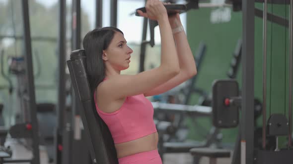 A Brunette Hispanic Woman in a Pink Suit Raises a Dumbbell to the Top While Training Her Shoulders alt