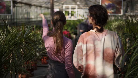 Back View of Young Female Florist Walking with a Client and Showing Her Different Plants Explaining alt