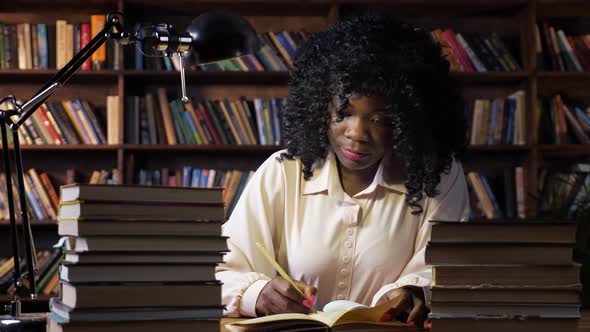 AfricanAmerican Lady Writes Near Stacks of Books at Table alt