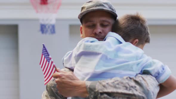 Happy caucasian male soldier hugging his smiling son holding flag in garden outside their house alt
