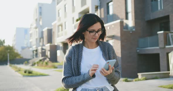 Young Caucasian Woman Using Mobile Phone During Walking Urban City at Sunny Windy Weather alt