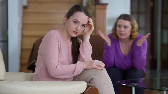 Overwhelmed Caucasian Young Woman Sitting at Home As Angry Mother Shouting Gesturing at Background alt