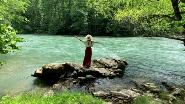 Young beautiful vintage woman in long skirt and straw hat dancing on stones near mountain river