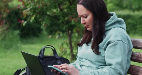 Brunette Woman Is Using Browsing Smartphone Sitting On Bench In Park Outdoors
