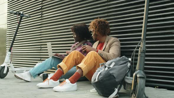 Black Man and Woman Sitting on Street, Using Laptop and Speaking alt