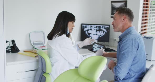 Biracial female dentist with face mask examining teeth of male patient at modern dental clinic alt