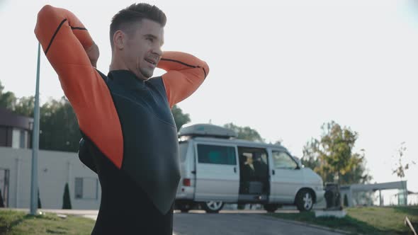 Man Puts on a Wetsuit on a Sunny Day with Van at the Background alt