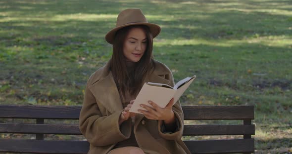 Cute Caucasian Female Student in Elegant Brown Clothes Sitting on the Bench and Turning Book Pages alt