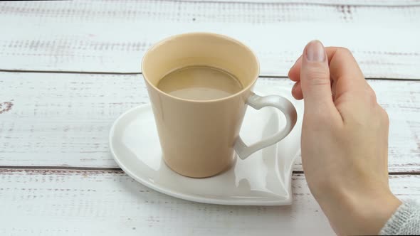 The Girl's Hand Takes a Cup of Coffee Close-up. Coffee Cup on a Saucer in the Shape of a Heart on a alt