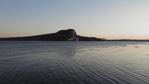 Flying away from shore towards a lone mountain across a lake at dawn AERIAL alt