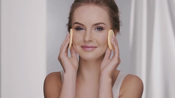 Happy Female Removing Make-up with Two Cotton Pads on Grey Background with Curtains alt