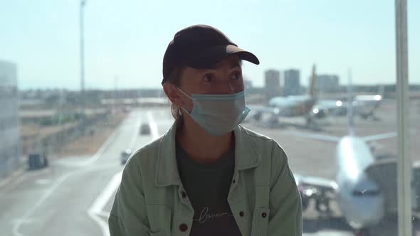 A Worried Woman in a Medical Mask at the Airport Sits Near a Window Against the Backdrop of alt