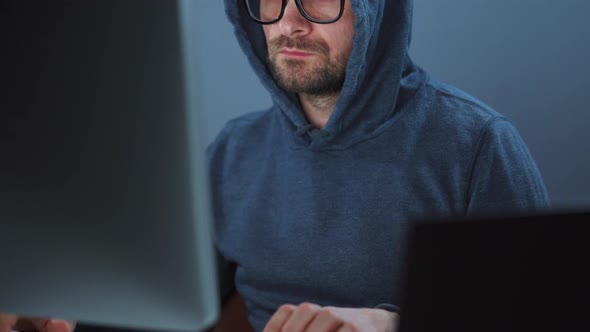 Male Hacker in the Hood and Glasses Working on a Computer in a Dark Office Room. Cybercrime Concept alt