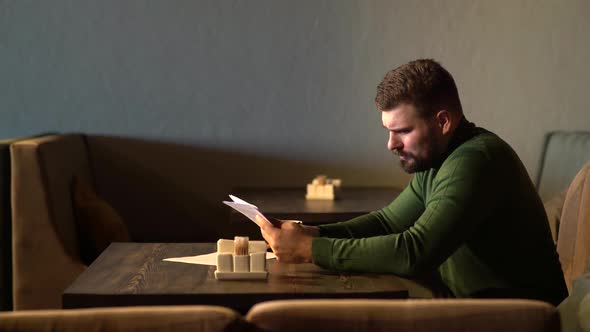 Young Attractive Bearded Hipster Man Sits in Cafe Reading Paper Documents