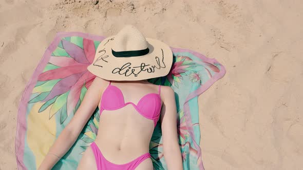 A Young Girl Lying on the Sand and Sunbathing Covering Her Face with a Big Hat alt