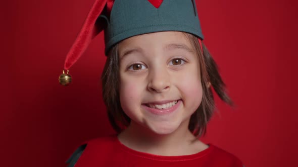 Baby Boy in a Green Suit and a Dwarf Hat is Indulging on Red Background for Christmas alt