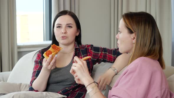 Two Girlfriend Girls Eat Pizza and Talk Sitting on a Couch in the Apartment