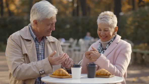 Senior Couple Disinfecting Hands with Sprayer Enjoying Snack At Outdoor Cafe alt