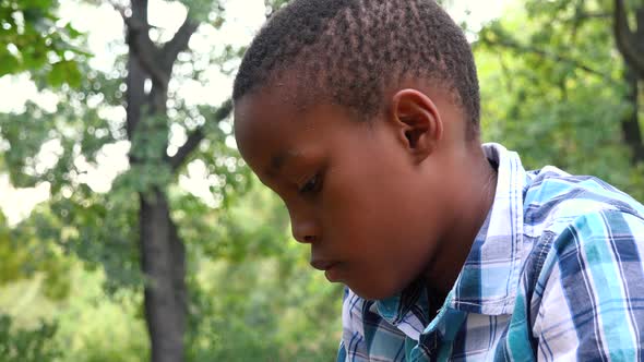 A Young Black Boy Sits on Grass in a Park and Works on a Smartphone - Closeup alt