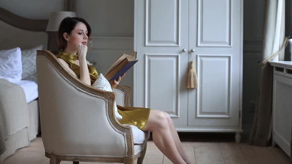 Young woman sits in armchair and reading book in vintage bedroom