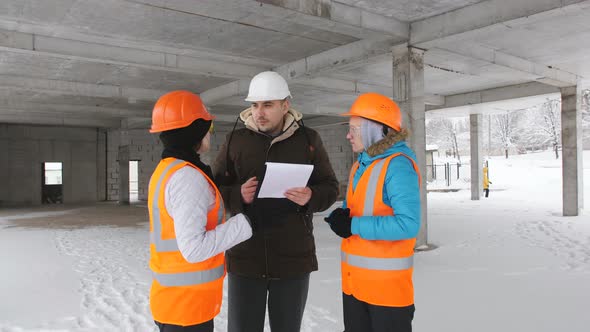 Two Female Engineers with a Folder and with a Helmet on Their Head Communicates with the Builder in