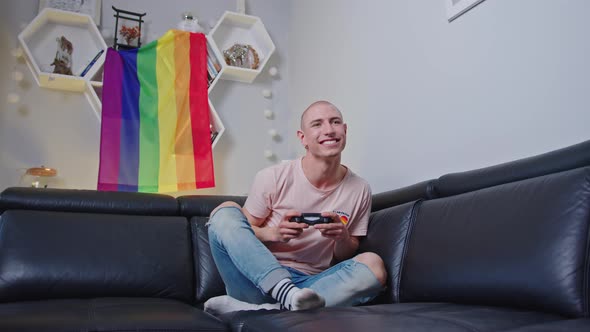 Bald Queer European Gay Activist Using Gamepad and Wining a Game While Sitting on Black Couch alt