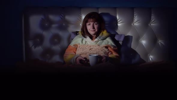 Woman Lies on the Bed at Night and Watches TV Switches Channels Drinks Tea and Smiles