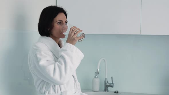 Thirsty smiling 35s woman in white bathrobe drinking water