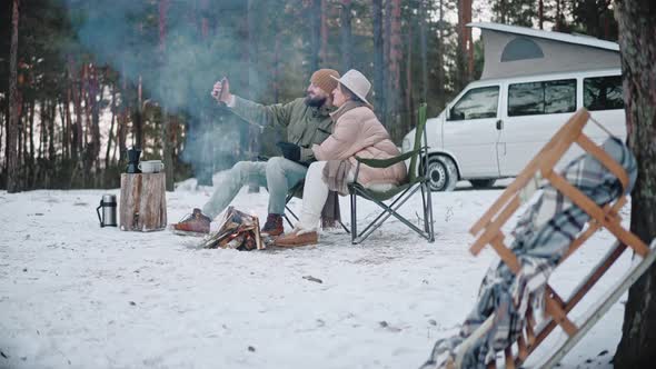 Couple of Travelers Sits By a Campfire and Takes Selfies in the Woods in Winter