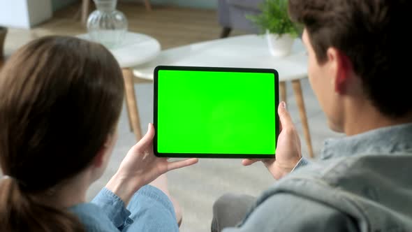 Over The Shoulder View Young Couple at Home Sitting on a Sofa Using With Green Mock-up Screen Tablet alt