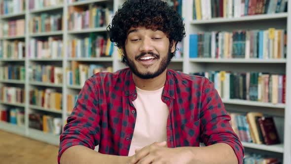 Positive Arabian or Indian University Stylish Student with Curly Hair Sits at a Table in the Library alt