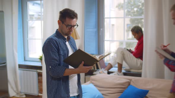 Young Guy Hipster Reading Textbook Walking in Dorm Room with Other Students Study alt