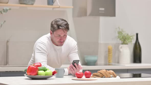Middle Aged Man Doing Video Call on Smartphone While Standing in Kitchen alt
