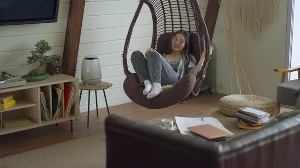 Wide Shot of Smiling Beautiful Asian Woman in Eyeglasses Sitting in Hanging Chair in Home Office alt