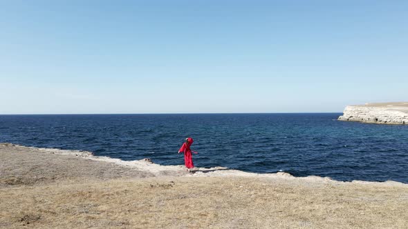 Brunette Woman in a Red Long Dress Stands on the Edge of a Cliff By the Sea alt
