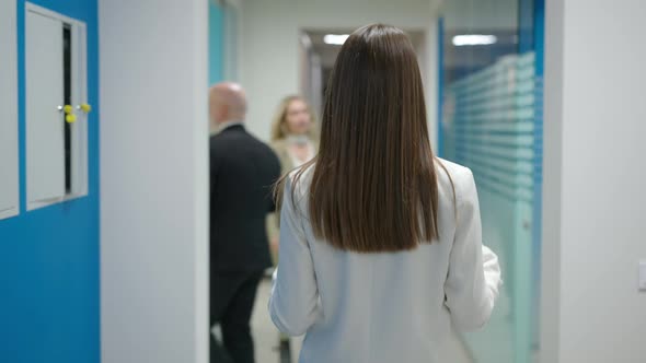 Back View of Slim Brunette Young Woman Walking in Office Hallway with Colleagues Passing alt