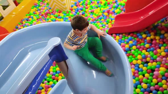 A Cheerful Preschooler Boy Descends From a Blue Slide Into the Pool with Balls in the Playroom alt