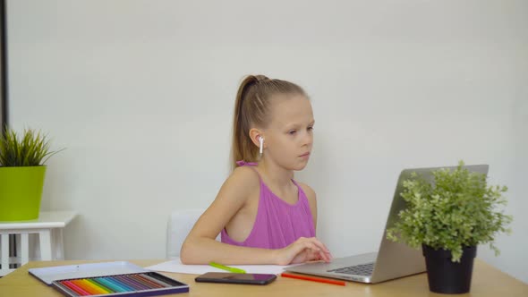 Serious Schoolgirl Sitting at Table with Laptop and Textbook and Doing Homework.