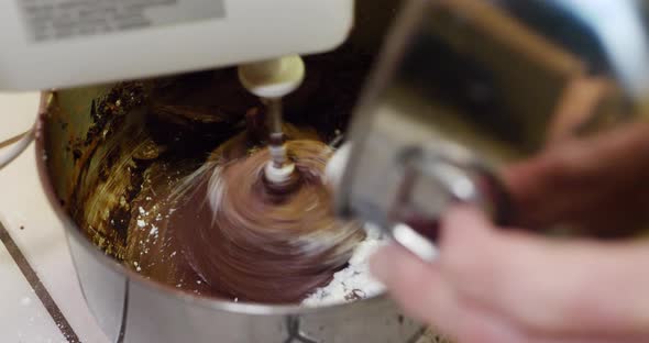 A chef in a kitchen using an electric mixer to make chocolate frosting for a cake in a metal mixing alt