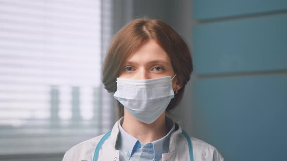 Red haired woman surgeon with blue eyes puts on disposable face mask and looks straight standing alt
